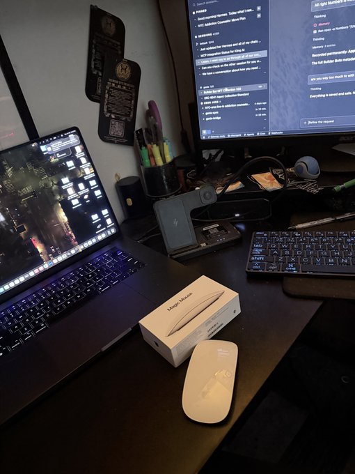 A desk setup featuring a laptop, external monitor, keyboard, and a boxed Apple Magic Mouse with the white mouse next to it.