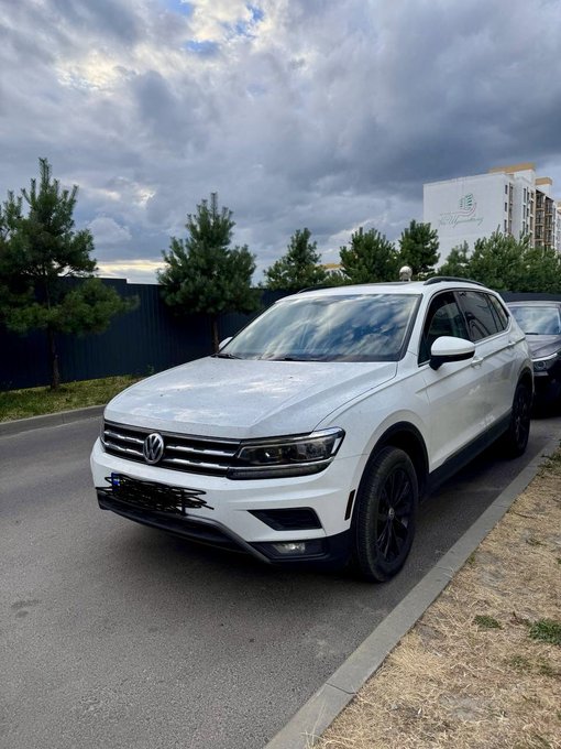 A white Volkswagen SUV is parked on a street with trees and apartment buildings visible in the background under a cloudy sky.