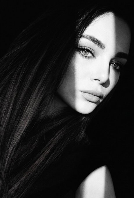 A high-contrast black and white portrait of a woman with long dark hair and striking eyes, lit dramatically against a dark background.