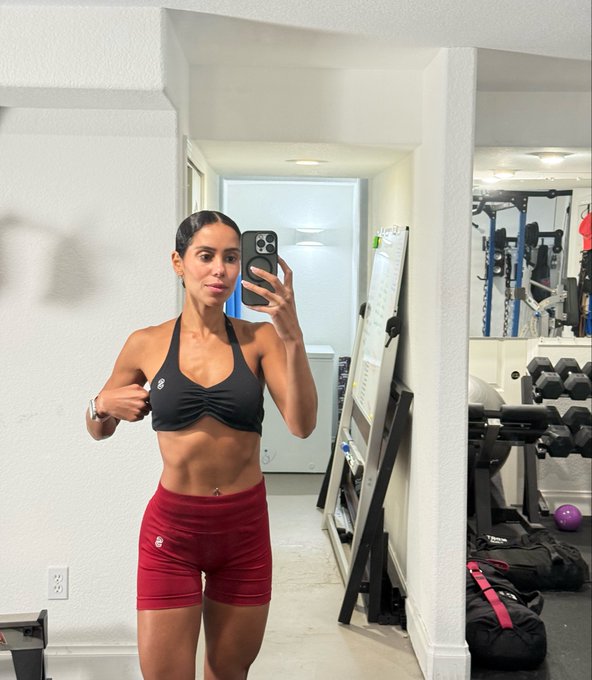 A woman with a toned body takes a mirror selfie in a gym, showing off her abs in a black sports bra and red shorts.