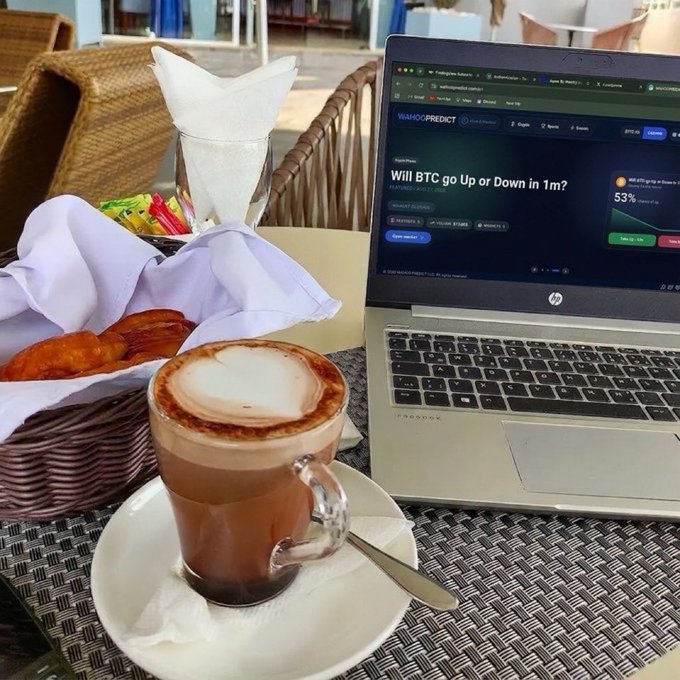 A glass cup of coffee with heart-shaped latte art sits on a saucer next to an open laptop displaying a 'WAGMI PREDICT' website asking 'Will BTC go Up or Down in 1m?', with a bread basket nearby.
