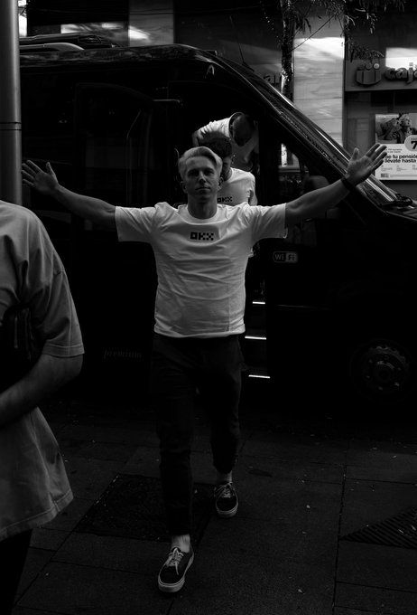 A black and white photograph of a person wearing an 'OKX' t-shirt standing with arms outstretched on a city street, with a dark van and another individual visible in the background.