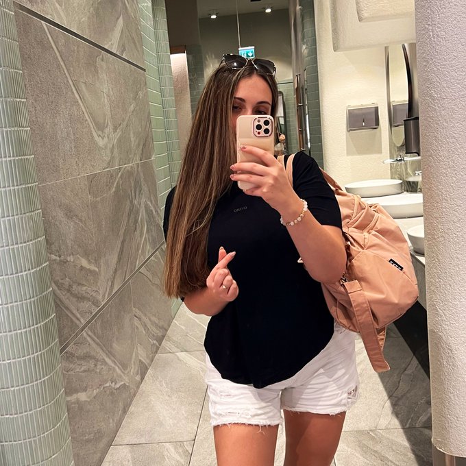 A woman with long brown hair takes a mirror selfie in a public restroom, wearing a black t-shirt, white denim shorts, and carrying a pink backpack.