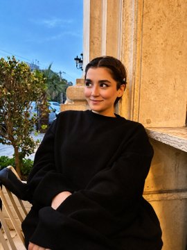 A woman with dark hair pulled back, wearing a black sweater, sits outdoors on a wooden bench leaning against a stone pillar.