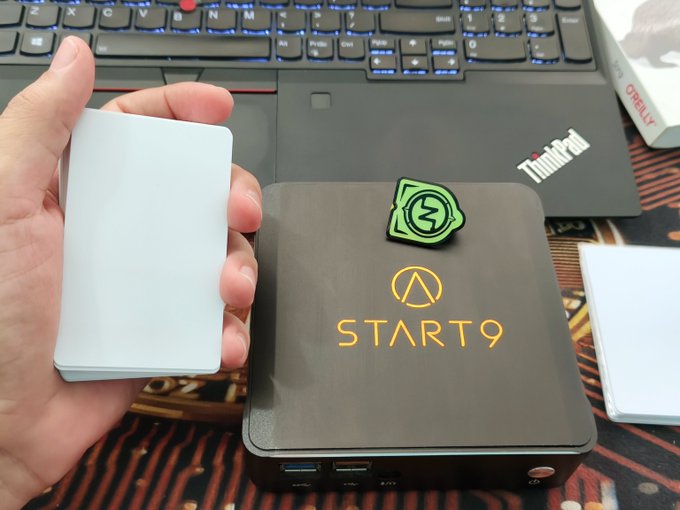 A hand holding a white device next to a black 'START9' box and a Lenovo ThinkPad laptop, with a green sticker resting on the box.