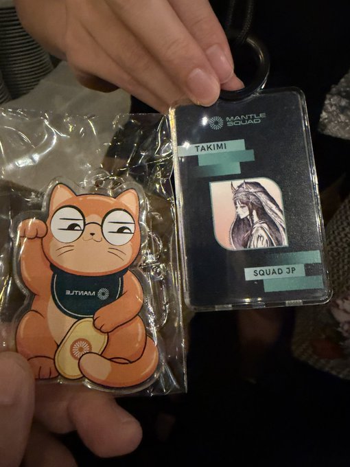 A person holds up exclusive Mantle Network swag, including a maneki-neko (lucky cat) mascot keychain and an ID badge for "TAKIMI" of "SQUAD JP," showcasing event memorabilia from the WebX conference.