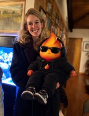 A smiling woman in a black coat holds up a small figure with a bright orange flame for a head, wearing black sunglasses and a black outfit with sneakers.