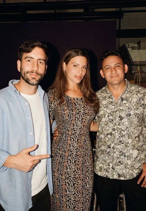 Three people pose together for a photo at an indoor event, with the man on the left making a hand gesture.