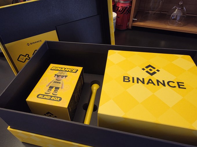 The image features a pair of glasses, likely for children or young people who need them. The glasses are placed inside a box with a yellow and black design, which has the word "binance" written on it. There's also a pen in the box along with the glasses.