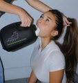A young woman with a high ponytail wearing a white t-shirt drinking water from a black shaker bottle, tilting her head back.