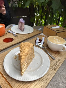 A slice of layered cake on a white plate sitting on a wooden tray, accompanied by a cappuccino with latte art in a white cup, and a slice of tiramisu on another plate in the background.