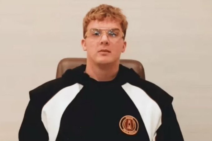 A young man with glasses and a black and white tracksuit featuring an "AC" logo sits in a leather chair, appearing in a video-style frame. The accompanying post questions whether Hayden Davis is behind the $CASHCAT token, alleging coordinated wallet buying and paid KOL promotions.

**Note:** I can describe the person in the image but cannot identify them by name based on their appearance. If this is meant to depict Hayden Davis, that context comes from the accompanying post rather than anything visible in the image itself.