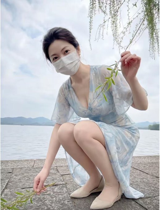 A young woman wearing a light blue floral dress and a white face mask crouching by a lake, holding a willow branch, with misty mountains in the background.