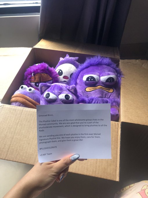 A hand holds a card addressed to 'Simonid Bimli' from the Monad community (#PLUSHCELEBRATE, MF Team) in front of a cardboard box filled with purple plushie toys, with the note explaining the plushies are part of the first-ever Monad Signature Plushie line.