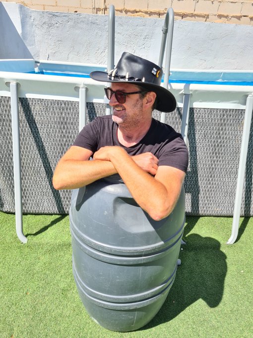 A man wearing sunglasses and a black cowboy hat smiling while sitting inside a large gray plastic barrel outdoors on artificial turf near a pool.