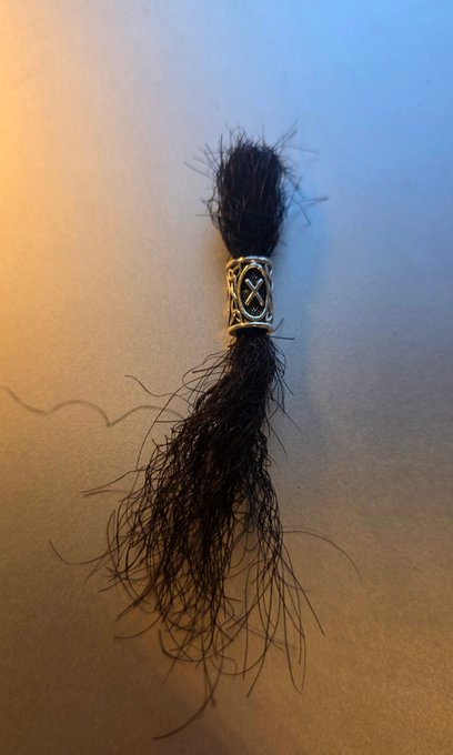 A bundle of dark hair wrapped with a decorative metal bead featuring crossed bones/X patterns, playfully serving as the "captain's iconic beard" being auctioned off for charity to fund the owner's erectile dysfunction treatment.