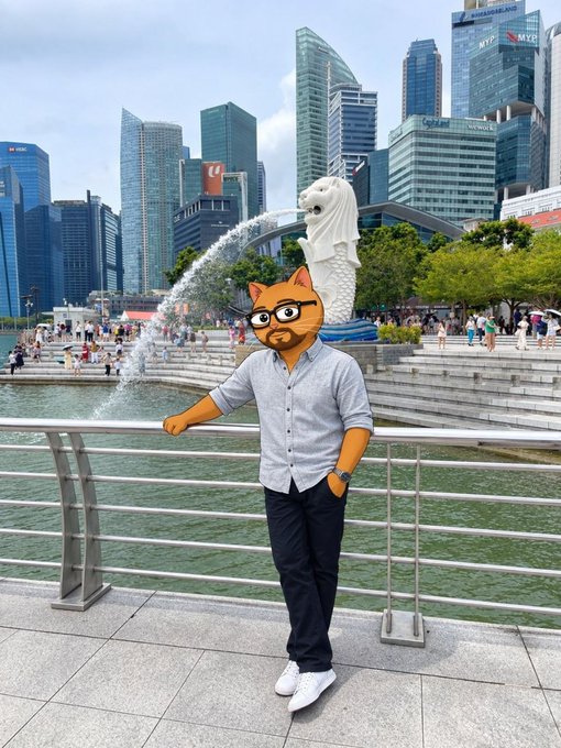 A man with an orange cartoon cat head, wearing a grey button-up shirt and dark pants, stands in front of the Merlion statue in Singapore with the city skyline behind him.