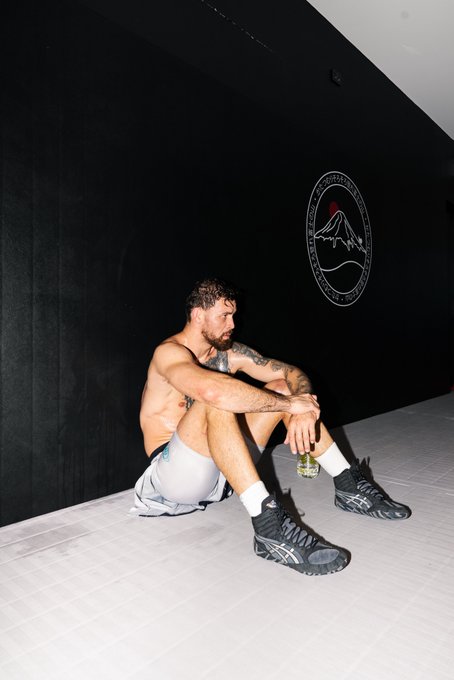 A shirtless, tattooed man in gray shorts and black ASICS sneakers sits on the mat of a black-walled training gym, gazing off to the side with a focused expression. The image reflects the intense preparation behind the post, capturing a moment of quiet concentration during a high-stakes training camp ahead of UFC 331.