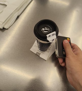 A first-person view of a hand using a smartphone to tap-to-pay at a payment terminal while holding a black reusable coffee cup on a metal counter.