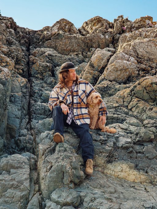 A man in a plaid flannel shirt and a bowler hat sits on rocky terrain next to a small dog, with both looking off to the side.
