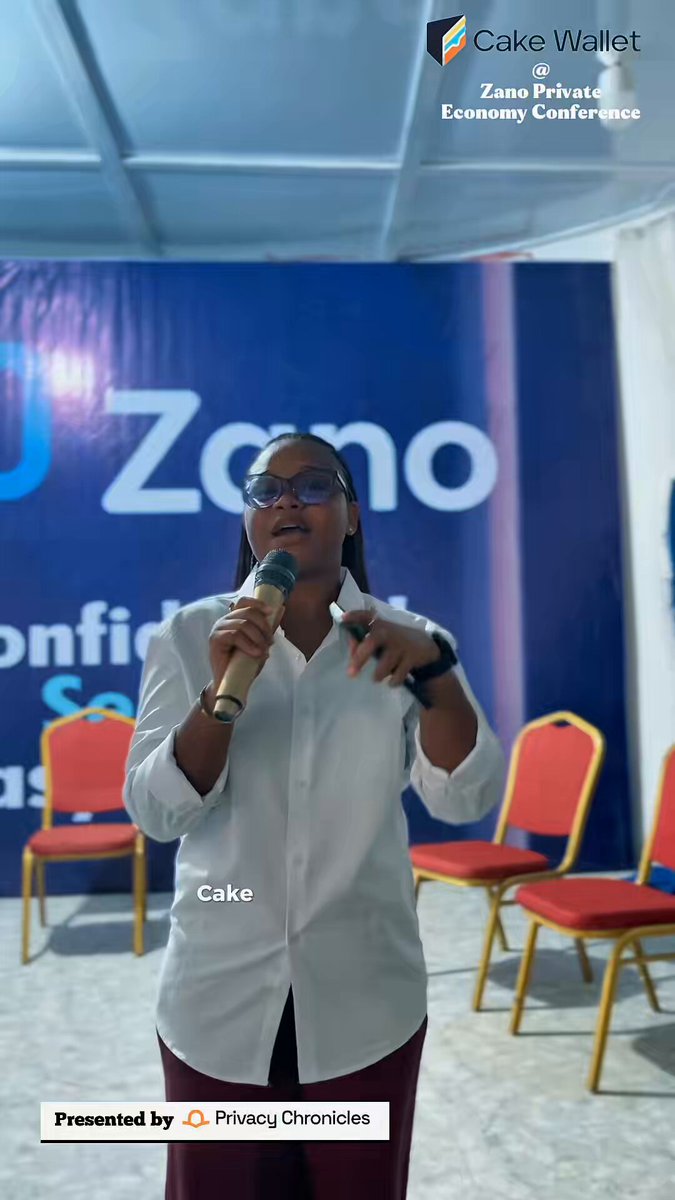 The image features a woman standing at a podium with a microphone, giving a speech or presentation. She appears to be wearing glasses and has her hands up, possibly emphasizing a point she's making. The setting seems to be an event or conference where people gather for discussions on various topics. There are several chairs in the background, indicating that this is likely a formal gathering or meeting space.