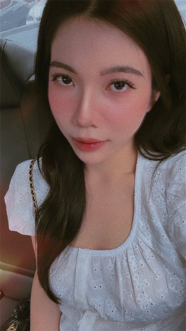 A selfie of a young woman with long dark hair, wearing a light blue embroidered top and a chain strap bag, taken inside a vehicle.