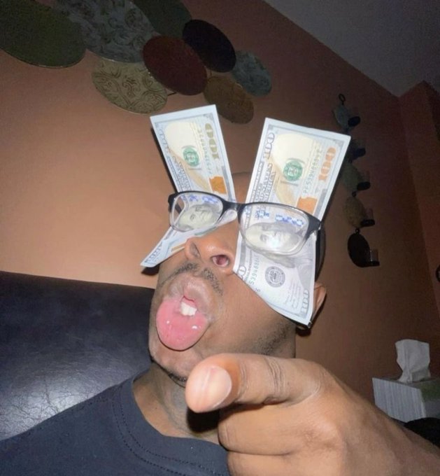 A man poses for a selfie with $100 bills folded over his glasses, sticking out his tongue and pointing at the camera.
