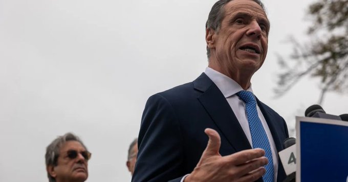 A man in a dark suit and blue tie, resembling Andrew Cuomo, speaking outdoors with microphones visible in the foreground.