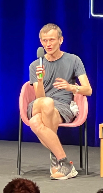 A man with short hair wearing a gray t-shirt and gray shorts, holding a microphone while seated in a pink chair on a stage with a blue curtain backdrop, appearing to speak at a panel or interview event.