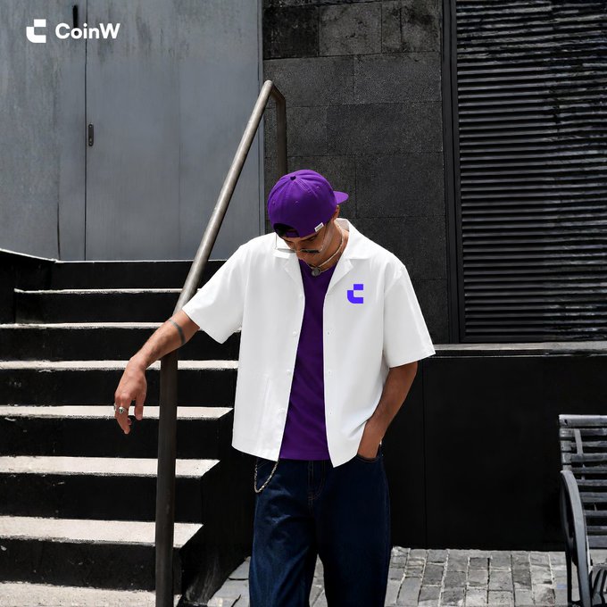 A model showcases a CoinW-branded streetwear look featuring a white button-up shirt with the purple "C" logo, paired with a purple t-shirt, matching cap, and dark jeans. The top-left "CoinW" watermark ties directly to the post's question about which piece from the collection fans would grab first—likely the bold purple cap or logo shirt as statement pieces.