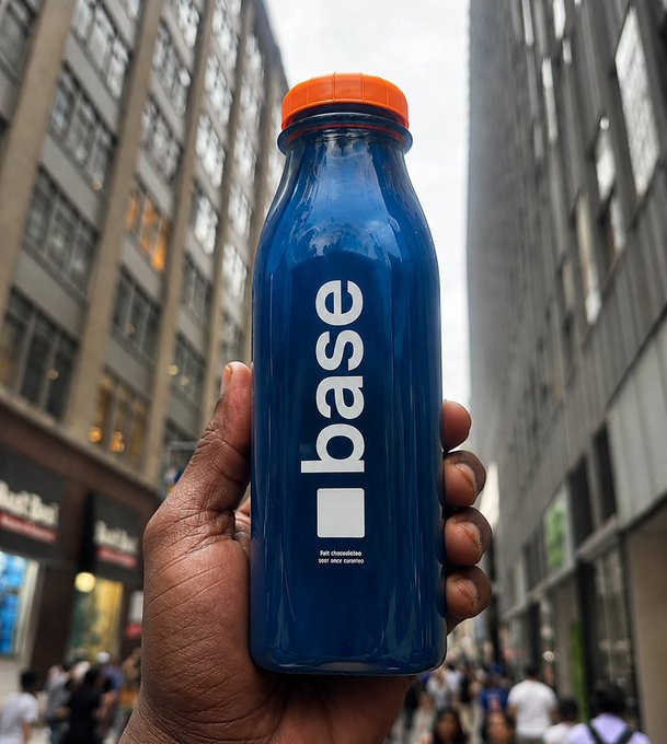 A hand holds a blue 'base' water bottle with an orange cap against a city street backdrop.