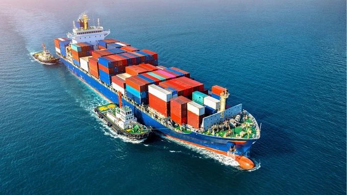An aerial view shows a large container ship loaded with colorful shipping containers being escorted by tugboats on the open ocean.