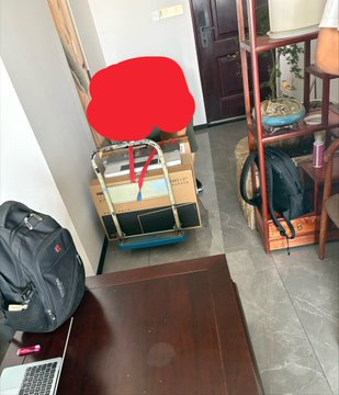 A cluttered indoor space with a backpack, luggage, a cardboard box, and a dark wooden table, with a red flower-shaped graphic covering part of the view.