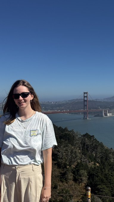 A woman wearing sunglasses and a tie-dye t-shirt stands in front of the Golden Gate Bridge on a clear, sunny day.