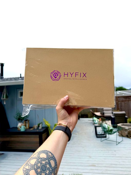 A hand holds up a sealed cardboard box branded "HYFIX SPATIAL INTELLIGENCE" against a backyard setting, representing the first $GEOD miner being shipped to a deployer through the Bounty Program.