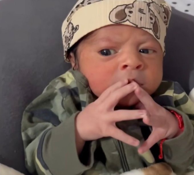 A close-up photo of a baby wearing a cartoon-patterned hat and camouflage jacket, with hands clasped together near their mouth.