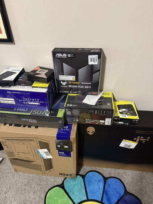 A collection of PC components is stacked on the floor, including an ASUS TUF Gaming B650M-Plus WiFi motherboard, NZXT H2 Flow case, MSI Pro B650 WiFi, a Wi-Fi 7 Pro box, Corsair Dominator Platinum RGB DDR5, and other parts.