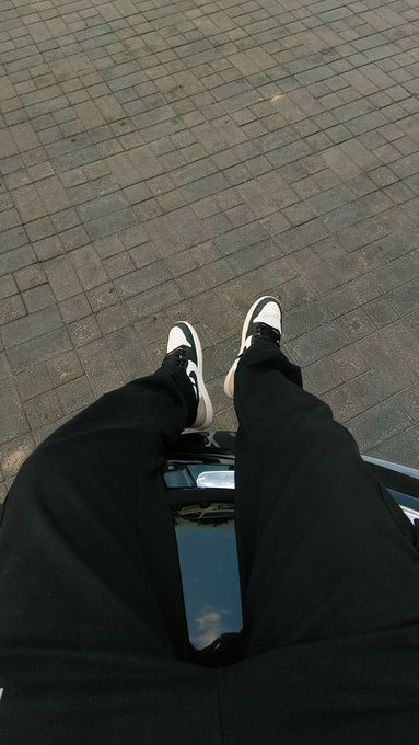 A first-person perspective looking down at someone wearing black pants and white-and-black sneakers, sitting with feet resting on a black surface on brick pavement.