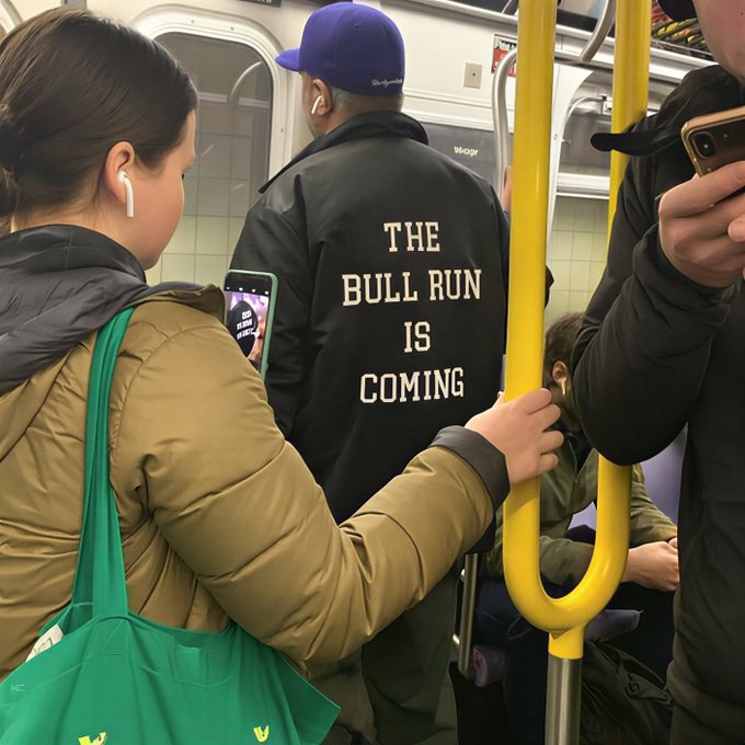 A man on the subway wears a black jacket emblazoned with "THE BULL RUN IS COMING" as a nearby woman holds up her phone, possibly photographing the timely message about the impending bull run.