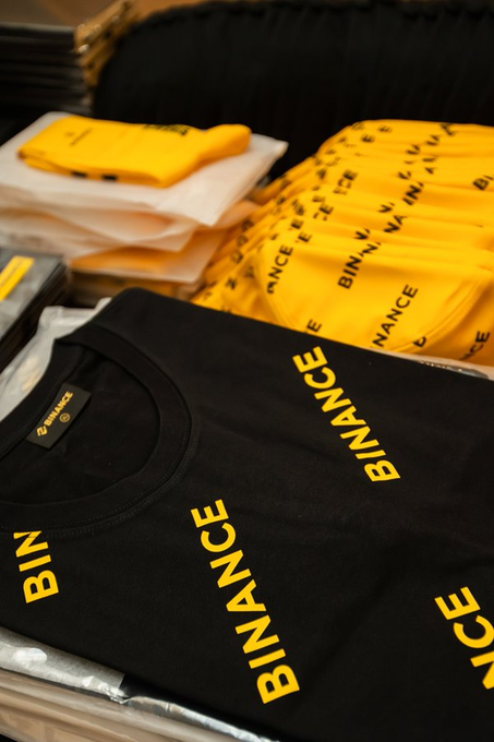 A display of folded Binance-branded merchandise, including black and yellow t-shirts with 'BINANCE' printed in yellow lettering.