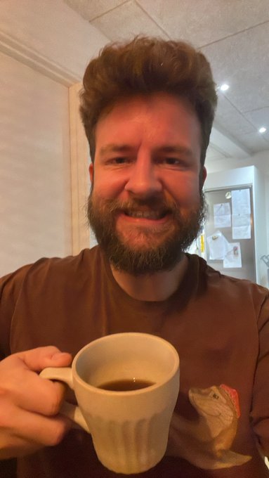 A smiling bearded man in a brown t-shirt holds a white mug of dark coffee indoors.