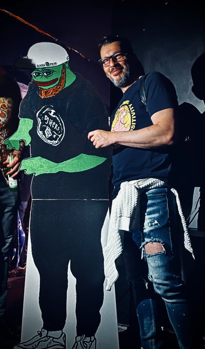 A man with glasses and a black t-shirt posing next to a cardboard cutout of a green cartoon character wearing a cap and sunglasses