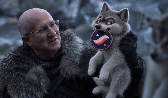 A man wearing glasses and a fur-collared coat holding up a cartoon-style wolf pup that has a striped emblem in its mouth, set against a snowy mountainous backdrop.