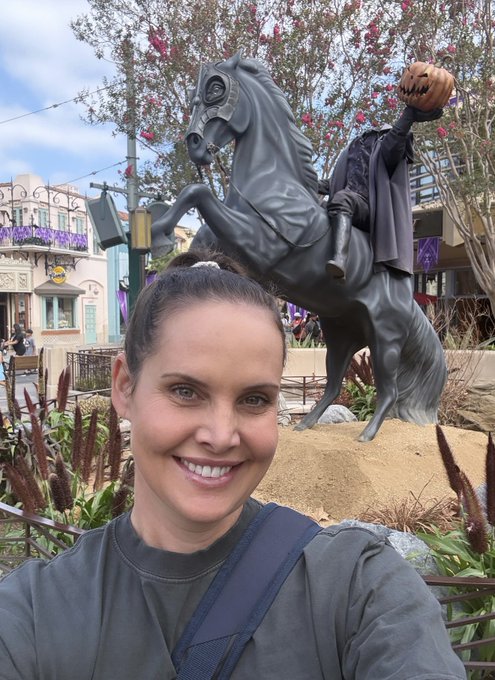 A woman smiles in front of a Headless Horseman statue holding a jack-o'-lantern at Disneyland, capturing the spooky spirit of Halloween time at the park.