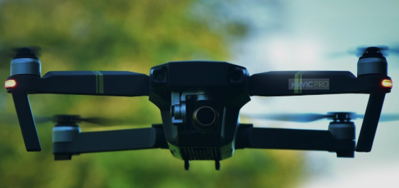 The image features a black drone flying through the air with its propellers spinning rapidly. The drone has four propellers attached to each wing, indicating that it is designed for low-altitude flight or hovering. The background of the image shows a blurred green landscape, which contrasts with the dark color of the drone and emphasizes its presence in the scene.