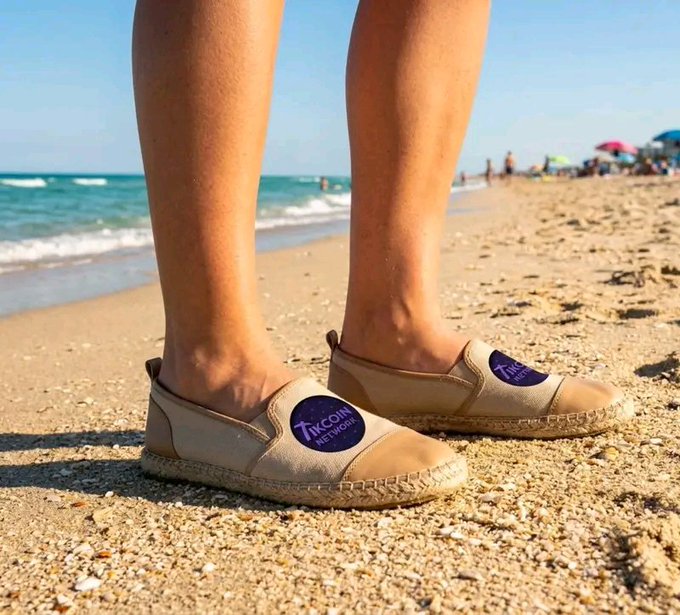 A person's legs stand on a sandy beach wearing beige espadrille shoes with purple circular labels reading 'ZKCOIN NETWORK'.