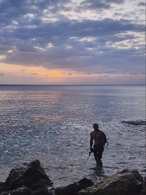 A scenic sunset view over the ocean, featuring the silhouette of a shirtless man holding a fishing spear while standing in shallow water near rocks.