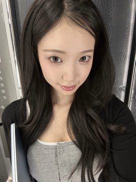 A young woman with long dark hair wearing a gray button-up top and black cardigan poses for a close-up selfie.