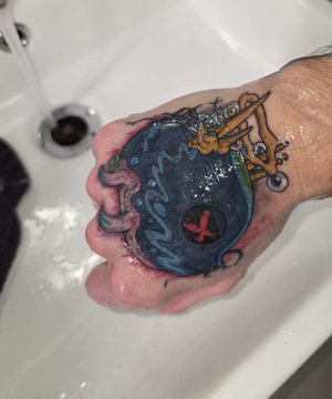 A fist with a colorful tattoo of a blue creature with an 'X' for an eye is held under running water in a sink.
