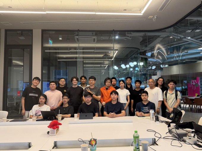 A group photo of about 18 young people gathered around a large conference table in a modern office space.